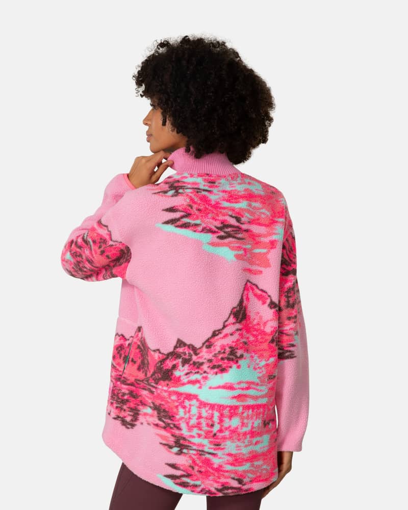 Back view of a woman wearing the pink Kari Traa Ruthie Midlayer with its mountain graphic and dropped back hem for coverage.