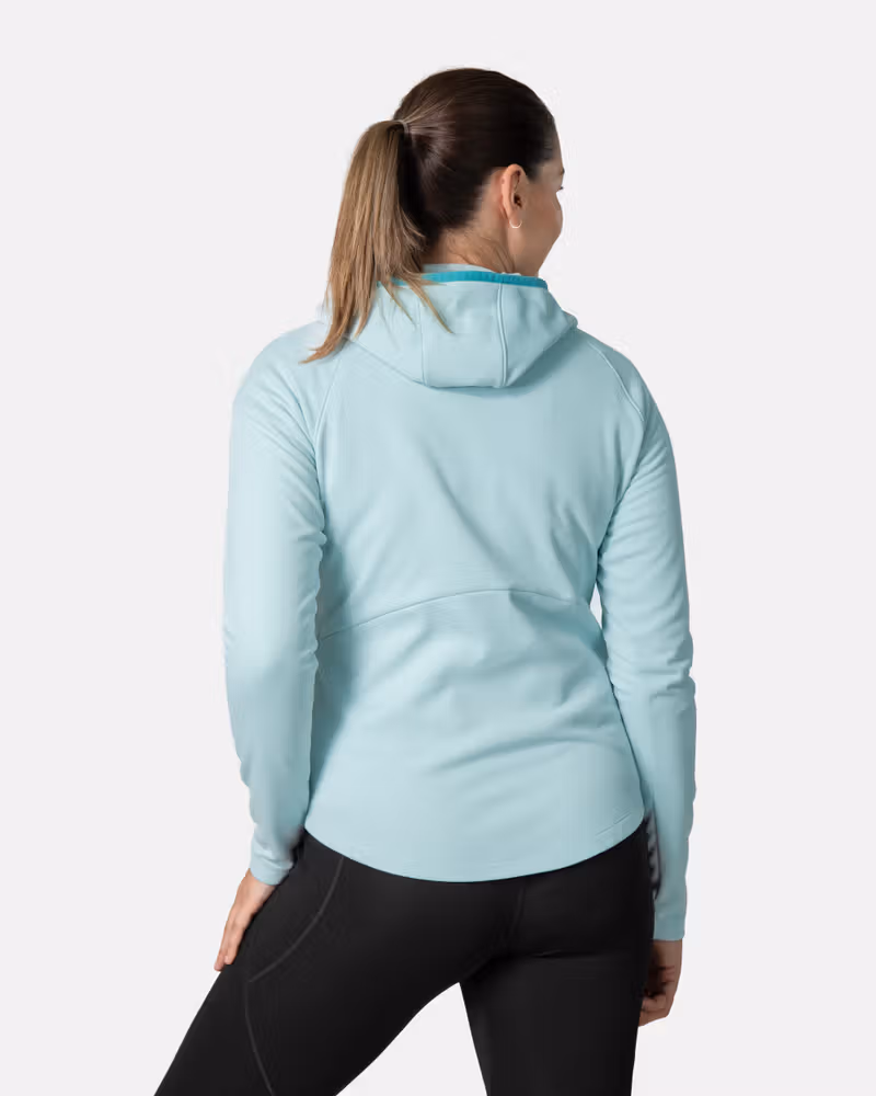 Back view of a woman in the light blue Kari Traa Nia Midlayer, showing the roomy hood and dropped back hem for coverage.