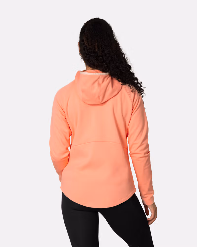 Back view of a woman in the peach Kari Traa Nia Midlayer, showing the roomy hood and dropped back hem for coverage.